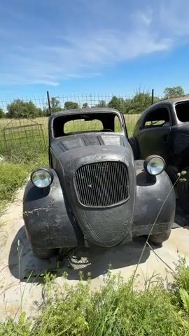 The video showcases two old, black cars parked outdoors on a sunny day. The cars appear to be in a state of disrepair, with no windshields and visible rust. The surroundings include green grass, weeds, a wire fence, and a few trees in the background under a clear blue sky with some clouds. The camera slowly pans across the cars.