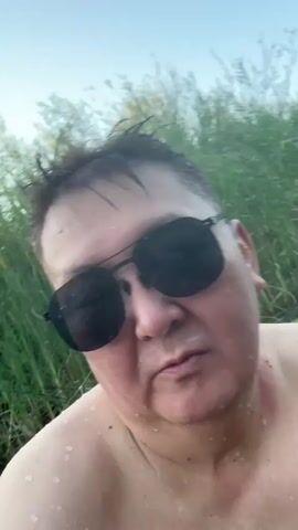A man wearing black sunglasses and no shirt is partially submerged in murky water, with tall green reeds visible behind him. He is looking directly at the camera. The water is rippling and appears to be flowing, with some splashes visible around his head and shoulders. The lighting suggests it is daytime, possibly late afternoon or early evening. The camera appears to be held close to the man, as it is a selfie-style shot.