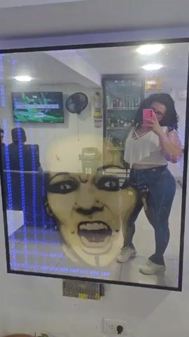 A woman with curly hair takes a selfie in a mirror. The mirror is rectangular and has colorful LED lights around the frame. There is an image of a person's face superimposed on the mirror. The woman is wearing a white shirt, patterned blue pants, and white sneakers. She is holding a pink phone and looking at the screen. In the background, there is a person sitting in a chair, a television, a fan, and a refrigerator filled with drinks. The room has white walls and a tiled floor.