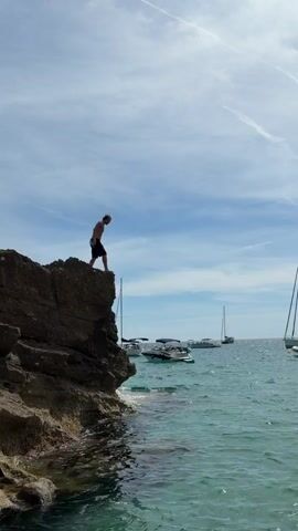 A person jumps off a cliff into the ocean. The person is wearing dark shorts and no shirt. The cliff is rocky and the ocean is clear and blue. Several boats are anchored in the background. The sky is blue with some white clouds. The jump and splash are captured in a short video clip.
