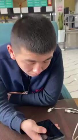 The video shows the interior of a space with a reception desk that reads "BURKUT" in large letters. There are also several blue upholstered booths with tables. Initially, a young person with a dark jacket and a red mark on their face is visible. Later, another man is seen sitting in one of the booths looking at his phone. The space has a modern aesthetic with patterned tile flooring and plants. There is also a television on the wall.