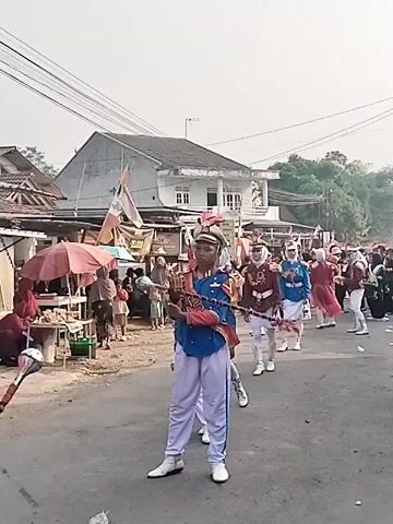 A marching band is parading down a street. The band members are wearing colorful uniforms, including blue jackets with gold trim, white pants, and white hats. The lead person in the band is holding a baton. The street is lined with spectators and there are several stalls selling goods. Buildings are visible in the background. The sky is overcast. The video is shot from a low angle, looking up at the marching band.