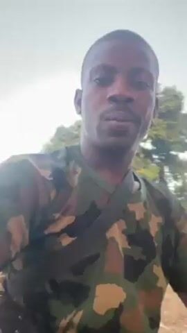 A man in a camouflage shirt is recording a selfie video. He is wearing a strap across his chest. He is outdoors with a building and trees in the background. He appears to be speaking but there is no audio.