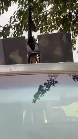 The video shows a static close-up of the top part of a military vehicle, possibly a pickup truck, under tree branches.  Mounted on top of the vehicle is a weapon system, likely a machine gun, with protective shields. The windshield is dirty and reflects the surrounding greenery, and the interior is dimly visible. The scene is outdoor and appears to be in a rural or semi-rural area.