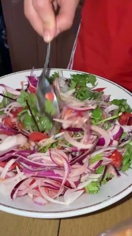 A close-up, slightly tilted shot shows a hand holding a spoon, mixing a salad in a white bowl. The salad contains thinly sliced red onions, chopped tomatoes, and fresh cilantro. The person mixing the salad is wearing a red shirt with white stripes on the sleeves. The bowl is placed on a wooden table.