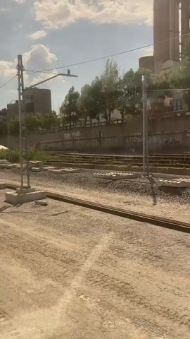 This media file captures a daytime scene, likely in the afternoon due to the strong shadows. The sky is blue with scattered white clouds, suggesting fair weather. The setting appears to be an outdoor construction or renovation site for a railway line.

The primary subjects are railway tracks, concrete sleepers, and power line poles characteristic of an electrified rail system. There are also construction materials such as concrete blocks and piles of gravel. In the background, industrial-looking buildings with circular structures are visible, along with trees and a long wall that has graffiti on it. A white tent or canopy is also present. A forklift is parked on the dusty ground in the foreground. There are no people clearly visible in the frame, although there might be distant figures in the background.

The activity taking place is the construction or maintenance of railway infrastructure. The presence of new concrete sleepers and the general state of the site indicate ongoing work.

Notable details include the presence of overhead power lines for trains, suggesting an active or soon-to-be-active electrified line. The dusty, rough terrain points to a work-in-progress environment. The graffiti on the background wall provides a cultural or urban element to the otherwise industrial and infrastructural scene. The time of day is indicated by the sun's position and the length of the shadows. The location cues (industrial buildings, potential for rail infrastructure) and the mention of Montcada i Reixac, Spain, place the scene within that specific geographical context, which is known for its railway junctions and industrial areas. No discernible text is clearly visible or legible within the media.