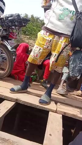 The video shows a group of people standing on a makeshift wooden bridge. The bridge is constructed of wooden planks with gaps between them, revealing a dark space underneath. One person is prominently featured, wearing yellow patterned shorts, dark socks, and sandals, and carrying a black bag. He is standing in the center of the frame. To the left, there is a parked motorcycle laden with items, and a person wearing a red garment is seated nearby. Other people are seated or standing around, some with luggage or bags. The environment appears to be rural, with foliage visible in the background under a bright, overcast sky. The camera moves slightly, showing the bridge from different angles.