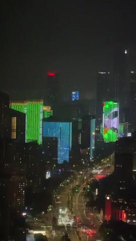 The video shows a nighttime aerial view of a city skyline with numerous tall buildings illuminated by colorful lights. A wide, busy road with a significant amount of car traffic runs through the center of the frame. The buildings are adorned with dynamic light displays, primarily in shades of green, blue, and multi-colored patterns, creating a vibrant and modern urban scene. The sky is dark and hazy, suggesting nighttime or possibly smog. The perspective is from a high vantage point, looking down and across the city.