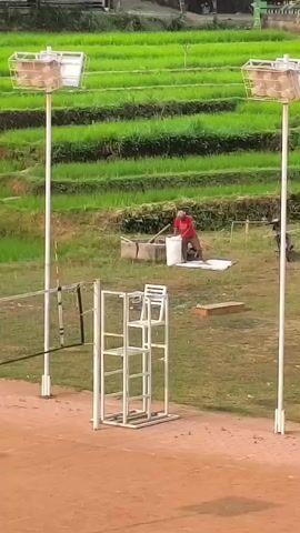 A person in a red shirt is working near a well in a rural setting. Behind the person is a lush green terraced rice paddy. In the foreground, there are volleyball court elements, including a net, umpire chairs, and tall light poles. The ground in the foreground is reddish-brown, suggesting a dirt court.