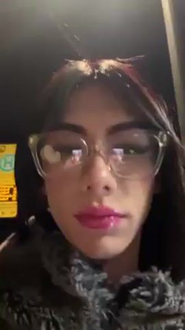 A close-up shot shows a person with long, dark hair and light skin wearing transparent, square-framed glasses. They have noticeable makeup, including pink lipstick and styled eyelashes. The person is wearing a dark, furry or textured coat. The background is dark, with some blurred lights and architectural details suggesting an outdoor urban environment at night. The camera is held at a slightly low angle, looking up at the person. The person is facing the camera and their expression is neutral.