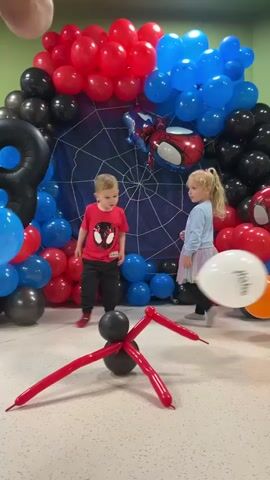 Two young children stand near an arch of red, blue, and black balloons in front of a backdrop resembling a spider web. One child, a boy, wears a red Spider-Man shirt, and the other, a girl, wears a light blue shirt and a skirt. There is a balloon spider on the floor. A person tosses a white balloon in the air. A third child, a boy, walks into the frame.