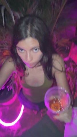 The video shows a person with long, dark hair in a tank top looking into the camera, followed by a shot of a crowd of people dancing at what appears to be an outdoor music event or party. The scene is lit with bright neon and colored lights, particularly pink, green, and purple. There are palm trees and other tropical plants visible in the background. Another person wearing a patterned shirt is seen briefly looking down, then looks up at the camera as the video ends. The atmosphere is lively and energetic.