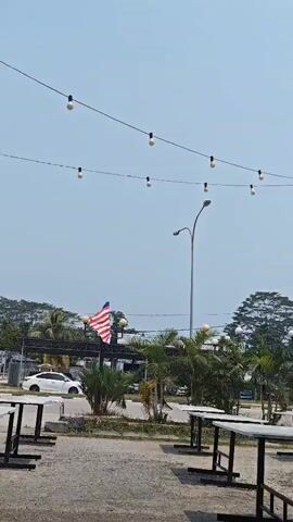 The video pans slowly across an outdoor seating area. Tables and chairs are arranged on a gravel surface. String lights are suspended overhead against a clear sky. A Malaysian flag is visible on a flagpole in the middle ground, flanked by palm trees and other vegetation. In the background, there are buildings, trees, and a street with a lamppost. A white car is visible driving on the street. A person walks across the frame from right to left in the foreground, passing the tables. The overall impression is of a casual outdoor dining or gathering space on a bright, clear day.