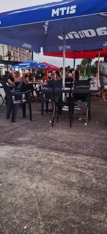 The video shows an outdoor seating area of a restaurant or bar. Several tables with chairs are visible, some occupied by people. Two large umbrellas, one blue and one red, provide shade. In the background, there are buildings, palm trees, and more people. The ground is paved. The scene appears to be set during the evening or late afternoon.