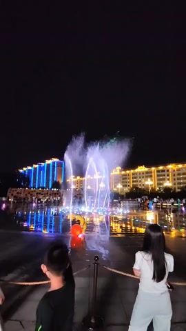 A nighttime aerial view captures a vibrant water fountain show. The fountain, illuminated with colorful lights, sprays water upwards in arcs. A large, modern building with blue and yellow lights stands behind the fountain, reflecting in the water on the ground. The area is paved and wet, suggesting recent activity. A group of people are watching the spectacle. In the foreground, two children are looking towards the fountain. A boy with glasses and a dark shirt is on the left, and a girl with long dark hair and a white top is on the right. They are separated by a thin rope barrier. The sky is dark and clear.