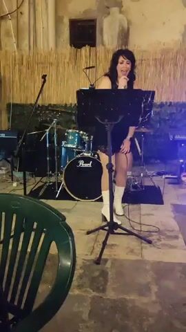 A female singer stands on a stage holding a microphone in her right hand. She is wearing a black dress and white boots. A music stand is in front of her. Behind her is a drum set and a keyboard. The background appears to be an outdoor setting with bamboo fencing and a brick wall. There is a green chair in the foreground. The video shows the singer performing.