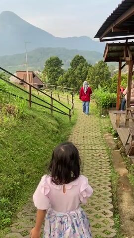 A young girl is seen from the back, running down a paved path. She's wearing a light pink dress with a colorful floral skirt. Ahead of her, a woman in a red sweater and hijab walks in the same direction. The path is bordered by green grass and a wooden fence on one side, and a wooden structure with a covered porch on the other. In the background, a mountain looms under a cloudy sky, with trees and houses scattered across the landscape. The girl pauses briefly to point to something off-camera before continuing down the path.