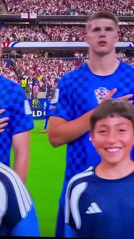 The video displays a scene from a soccer match, likely during the pre-game national anthem. The focus is on several Croatian soccer players in their blue and white checkered uniforms, standing with their hands on their chests. Alongside them, or in front of them, are several children, also wearing blue and white Adidas tracksuits, some smiling at the camera. The background shows a packed stadium with spectators wearing red and white attire. The field is green. The Croatian flag is visible on the players' jerseys and also on a flag in the background. The overall atmosphere appears to be formal and patriotic.