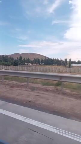 A car passenger's view of a rural landscape from a moving vehicle. The scene includes a highway guardrail in the foreground, with a dusty shoulder to its left. Beyond the guardrail is a fenced-off area with a few scattered trees and buildings. Further in the distance, a line of tall trees borders a sparsely vegetated hillside. The sky is blue with scattered clouds, and the overall impression is one of a sunny, clear day. The video appears to be shot during daylight hours.