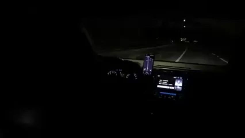 A night drive is shown from the perspective of the driver's seat of a car. The road ahead is illuminated by the car's headlights, showing a multilane highway with guardrails on either side. The car's dashboard is visible, including the steering wheel, instrument panel, and a center console with a touchscreen display. A phone is mounted on the dashboard to the right of the steering wheel. Music is playing. Traffic lights are visible in the distance.