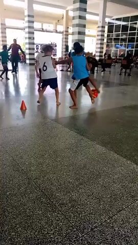 The video shows an indoor space with a speckled floor and tall, striped columns. In the foreground, four children are playing with a small orange ball. Two boys, one in a blue tank top and black shorts, the other in a white t-shirt and blue shorts, are actively engaged with the ball, appearing to be kicking it back and forth. To the left, another child in a white shirt and blue shorts watches, and further left, another child in a white shirt and blue shorts runs away from the ball. In the background, several adults and children are seated at tables, and a person in a red vest stands on the far left. The overall atmosphere seems to be that of a recreational activity or game.