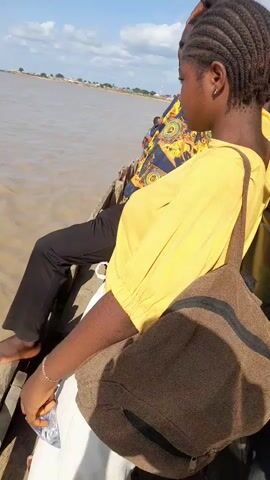 A video shows people sitting on the edge of a boat. The water is brown and choppy, and there is a shoreline in the distance with trees and buildings. The sky is blue with some clouds. The people are dressed in casual clothing. One person is wearing a yellow shirt and has a brown bag on their shoulder. Another person is wearing black pants and has their feet dangling in the water. The video is filmed from a close-up perspective, focusing on the people and the boat.