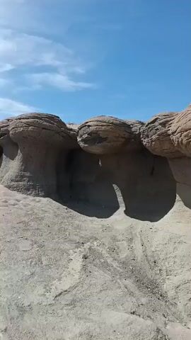 The video pans across a barren desert landscape with rock formations. In the foreground, peculiar mushroom-shaped rocks are visible, sculpted by erosion. The sky is a clear blue. A man in an orange shirt and dark shorts is seen walking along a dusty path beside the rock formations. He pauses to touch one of the rocks before continuing his walk along the edge of a cliff overlooking a valley. The terrain is dry and rocky with sparse vegetation.