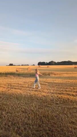 A young person with blonde hair, wearing a pink top and blue jeans, is running through a golden field of dried grass. Several large hay bales are scattered across the field. The sky is a clear light blue with a few wispy clouds. The sun is low in the sky, casting a warm, golden light over the scene. The person is moving from left to right across the frame, with their arms outstretched. Trees and more hay bales are visible in the distance.