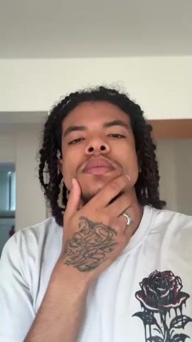 A person with dark, braided hair and tattoos on their hand and neck is in a room, looking at the camera. The person wears a white t-shirt with a rose graphic. The person touches their chin, then their neck, then covers their mouth with their hand, and finally raises their middle finger to the camera. The room has light-colored walls, a wooden floor, and a painting on the wall.