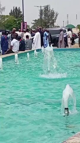 The video captures an outdoor scene in Unguwan Sanusi, Nigeria, featuring a large, rectangular fountain with turquoise-colored water in the foreground. Several water jets spray upwards from the fountain, creating a dynamic visual. In the background, a crowd of people, predominantly adults, is gathered around the fountain's edge. Some individuals are seated on the low wall of the fountain, while others stand, observing the surroundings or interacting.

The people exhibit diverse attire, including traditional robes (like a white agbada worn by one man prominently walking in the mid-ground) and more casual Western clothing. Behind the crowd, trees, streetlights, and a dark SUV are visible, suggesting a public space like a park or plaza. The bright lighting indicates it is daytime, likely sunny or partly cloudy. The overall atmosphere appears calm, suggesting a public gathering or casual observation rather than a specific event. A blurry purple banner with indistinct text is visible in the far background.