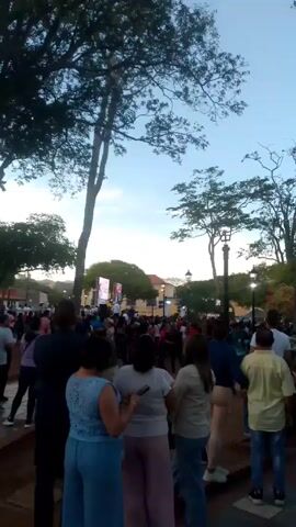 The video shows a large crowd of people gathered in a public square. In the background, there is a stage with two large screens displaying images. A few individuals appear to be on the stage. The crowd is mostly composed of people standing and watching the stage. Tall trees and street lamps surround the square. The sky is visible in the background, appearing blue with some cloud cover. The video appears to be taken from within the crowd, looking towards the stage.