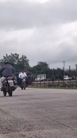 A man rides a motorcycle loaded with several stacked plastic crates. The motorcycle is on an asphalt road, and the man is wearing a white shirt and dark pants. In the background, there are trees, utility poles, and some vehicles. The sky is overcast with clouds.