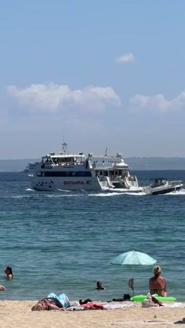 The video shows a bright, sunny day at a beach. In the foreground, people are relaxing on the sand and in the shallow water. A beach umbrella provides shade for one person on the sand. In the middle ground, several people are swimming or playing in the clear blue water. In the background, a large white ferry named "BRITANNIA JET" is sailing on the ocean, leaving a wake behind it. Another smaller boat is visible behind the ferry. The sky is blue with a few scattered white clouds. The water is calm and appears to be near a coastline.