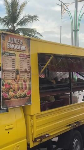 The video opens with a close-up shot of the side of a bright yellow food truck selling juices and smoothies. A menu board is visible on the side of the truck, displaying prices in RMG and listing various fruit juice and smoothie options. The truck is parked on an asphalt surface, likely a street or market area, with a palm tree visible in the background. The camera then pans to the right, revealing more of the market scene. A series of large, round, white LED lights are strung across wires, casting a soft glow. Several other food trucks and stalls are visible in the background, with people walking between them. Some of the trucks appear to be selling street food. The sky is overcast with some hints of blue peeking through. In the foreground, a table displays several large dispensers filled with orange-colored juice. Various fruits, including pineapples and oranges, are also displayed on the table. The video captures a bustling outdoor market atmosphere, with a focus on a juice and smoothie stand. Several individuals are seen walking through the market, some of whom are interacting with the vendors or browsing the stalls. The overall impression is of a lively street market on a cloudy day.