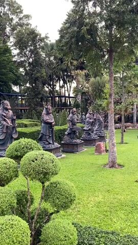 The video displays a well-manicured garden with several large, dark statues of robed figures standing in a line. The statues are positioned in front of meticulously trimmed hedges, creating a formal garden setting. In the foreground, there are several topiary bushes shaped into rounded forms, adding to the artistic landscaping. Behind the statues, lush green grass extends, and beyond that, a variety of trees, including palm trees and taller deciduous trees, form a natural backdrop. The sky appears overcast, suggesting a cloudy day. In the far background, hints of structures, possibly part of a larger complex, are visible through the trees. The scene is peaceful and orderly, highlighting the statues and the surrounding botanical arrangement.