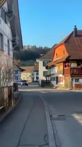 The scene is a quiet street in a European village with traditional architecture. The buildings are a mix of white and wooden-framed structures with red-tiled roofs, typical of Swiss or German villages. The street is asphalt with a concrete curb, showing slight signs of wear. The sky is clear and blue, indicating a sunny day. A black SUV is driving slowly down the street. There is a small hill with trees visible in the background. The overall atmosphere is peaceful and serene.