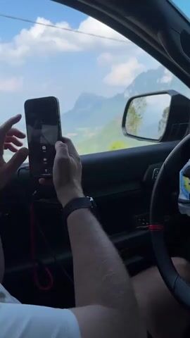 The video shows the view from the passenger seat of a car traveling through a mountainous landscape. The passenger is holding a phone and appears to be looking at images. The mountain range is lush with greenery, and the sky is blue with scattered clouds. The rearview mirror offers a reverse perspective of the road and scenery. The car's interior is black with red accents on the steering wheel. The passenger is wearing a white shirt and a dark-colored watch. There is music playing in the background.