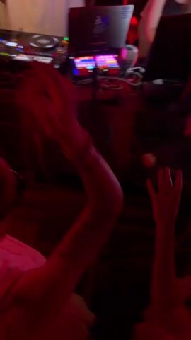 This media file captures a vibrant scene inside a nightclub, likely in Sofia, Bulgaria. The setting is characterized by a DJ booth with two turntables and a laptop, illuminated by pulsating purple and pink lights. The DJ, dressed in a white shirt, is actively engaged in mixing music, indicated by their hands on the equipment.

The foreground shows the backs of several individuals enjoying the music and atmosphere. One person in the center is wearing a white top and has their hair in a braid, with a glimpse of sunglasses. Another person to their right, also in a white top, appears to be dancing. The overall mood is energetic and celebratory, with the dynamic lighting contributing to a lively ambiance.

Notable details include the digital interfaces on the DJ equipment and the laptop screen, suggesting a modern setup for the performance. The reflection on the dark, possibly marble-patterned, surface of the DJ booth adds depth to the visual. While no specific time of day is evident, the club setting implies it's nighttime. The emotion conveyed is one of enjoyment and immersion in the music and social experience. No legible text is visible in the media.