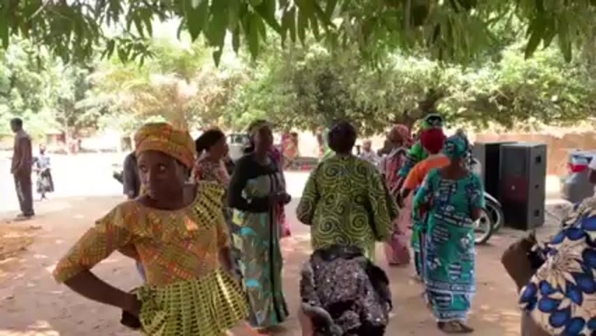 The video shows a group of people dancing outdoors under the shade of trees. Most of the dancers appear to be women, dressed in colorful patterned outfits and headwraps. Some people are standing around watching the dancers. A speaker is visible near the edge of the dancing area. The ground appears to be sandy or dirt.