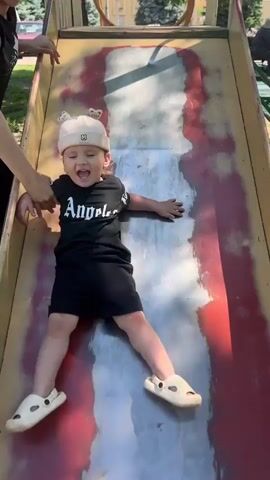 A young child wearing a white hat with animal ears and a black t-shirt with "Angeles" written on it, and black shorts, is sliding down a metal slide on a playground. The child is holding onto the sides of the slide. A person's hand is visible on the left side of the frame, holding the child's arm. The slide appears worn and has red and white markings. The ground around the slide is covered in dirt and leaves. Sunlight is filtering through trees, casting shadows on the slide.