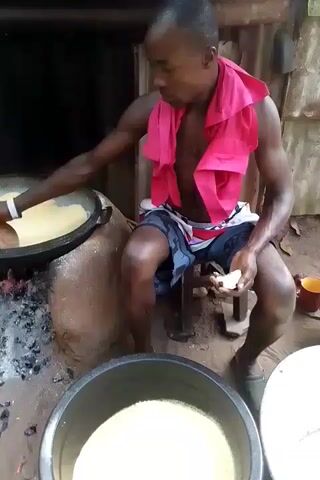 A man with short dark hair, wearing patterned shorts, a pink shirt draped around his neck, and sandals is seated on a small wooden stool, stirring the contents of a large black wok-like pan. The pan is perched on a makeshift stove built from clay or earth, with a fire burning underneath. The contents of the pan appear to be a light tan color. In the foreground, there is a black plastic tub filled with the same light tan substance. Another white tub is partially visible to the right. The man is holding a small white lump in his hand. He is looking down and concentrating on the pan as he stirs. The setting appears to be outdoors or in a simple structure with corrugated metal walls visible.