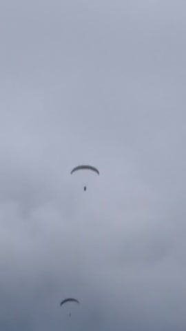 The video shows two paragliders in the sky. The paragliders are in the distance and appear to be high up. The sky is cloudy and overcast. The video pans down to reveal a rocky outcrop in the foreground, with some greenery on top. In the background, there are trees on a hillside, and a building is visible on the left side of the frame.