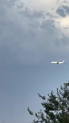 A commercial airplane is shown flying across a gray, cloudy sky. The plane is white with a blue tail. In the foreground, the tops of some buildings and trees are visible. The lighting suggests it is daytime, possibly before or after a storm.