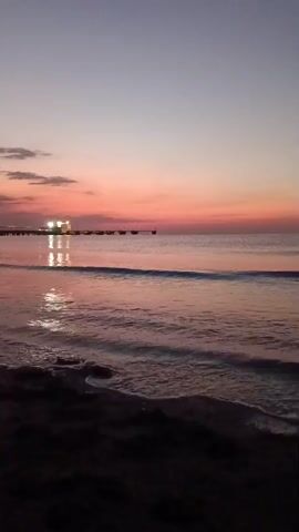 The video shows a calm ocean with gentle waves lapping against a sandy shore. In the distance, a pier with lights stretches out into the water. The sky is a gradient of soft pinks, purples, and blues, indicating either sunrise or sunset. The overall atmosphere is serene and peaceful.