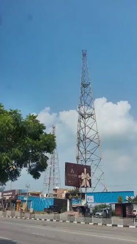 The video shows a street scene with a cell tower in the background. A red truck with a tarped cargo bed is driving on the road. There are other vehicles and buildings visible. The sky is blue with some clouds. The footage appears to be filmed from a moving vehicle.