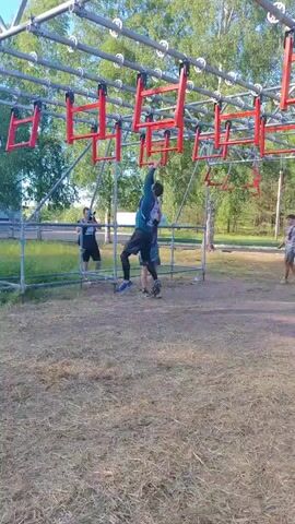 A person is traversing an obstacle course, swinging from red handles suspended from a metal frame. The person is wearing a dark blue and green shirt and black pants. They are in mid-swing, with their body angled and one leg bent. Another person, wearing a dark shirt and shorts, stands below and to the left, observing. To the right, another person in a dark shirt and shorts walks away. The obstacle course is set up outdoors on a grassy, dirt-covered area, with trees and foliage visible in the background under a clear sky. Scaffolding supports the structure. The timestamp is 00:00-00:03.