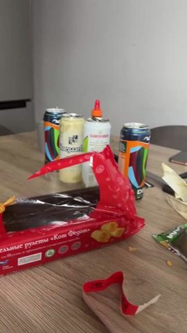 The video shows a messy wooden table with various items scattered across it. There are opened packages of chips and cookies, several cans of energy drinks and beer, a bottle of water, and multiple glasses, some with liquid in them. There are also mugs, a cutting board with cheese, a phone, and what appears to be packaged food. The general impression is one of disarray and possible remnants of a gathering or party. The background is a plain white wall.