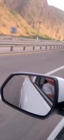 The video begins with a view of a highway with a mountain range in the background. A white truck is seen driving on the highway. The camera is positioned inside a vehicle, as indicated by the side-view mirror visible in the foreground. The scene is captured during daylight hours, with the sun illuminating the landscape. The road appears to be well-maintained, with clear lane markings and a guardrail separating the lanes. A few utility poles are visible along the side of the road. The video lasts for approximately 5 seconds, showing the truck passing by and the vehicle continuing its journey on the highway.
