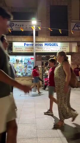 A medium shot shows a street scene at night. In the foreground, several people are walking. On the left, a couple walks away from the camera, the woman in black pants and a white top, the man in khaki shorts and a dark t-shirt. To their right, a woman with blonde hair in a red top and white shorts walks toward the camera, followed by another person partially visible. In the background, a store with a sign that reads "Ferreterías MIGUEL" is visible. The store appears to be a hardware store, with displays of various items visible through the window. More people are seen walking in front of the store. The street is paved with cobblestones, and streetlights illuminate the scene. The overall atmosphere is that of a bustling urban environment at night.