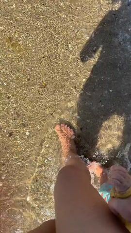 The video shows a first-person perspective of someone walking on a beach in Sérignan, France. The sandy shore is wet and reflective from the receding tide, with small shells and pebbles scattered across the surface. The sun is bright, casting a long shadow of the person onto the sand. The person's bare feet are visible, stepping into the shallow water and then back onto the sand. A young child's hand, wearing a yellow bracelet, is also visible in the frame, holding onto the person's leg. The overall mood is peaceful and relaxed, suggesting a pleasant day at the beach. The time of day appears to be daytime, with bright sunlight and clear weather.